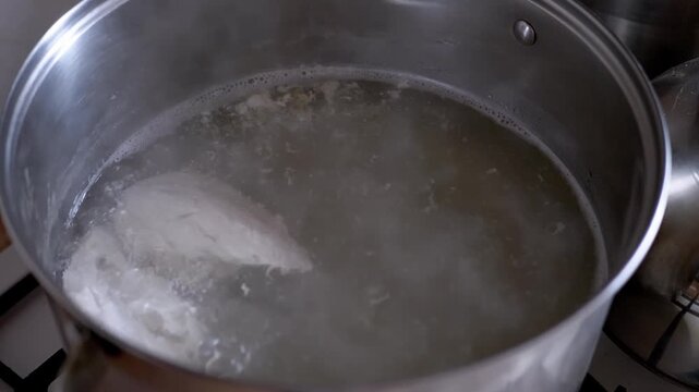 Fresh chicken breasts boil in bubbling water inside a stainless steel pot on a kitchen stove. Concept of healthy homemade meal preparation, dietary protein cooking, and traditional soup.