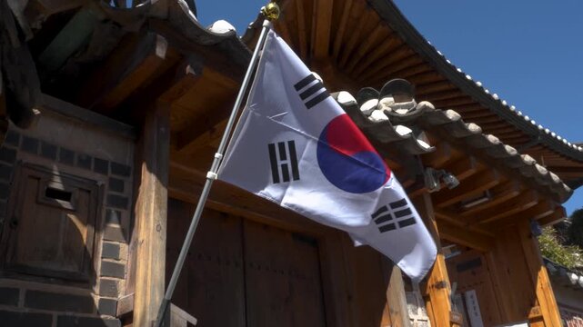 South Korean national flag Taegukgi waving on a pole in front of a traditional Korean hanok wooden architecture with tiled roof under a clear blue sky in Bukchon Hanok Village. Seoul, South Korea