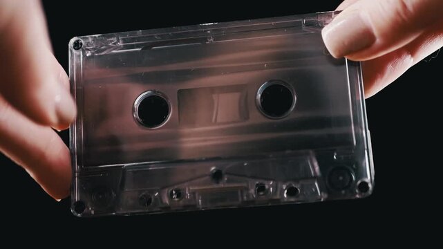 Hands gently hold and examine a clear analog audio cassette tape against a solid black background. Concept of vintage music technology, retro nostalgia, and classic media formats.