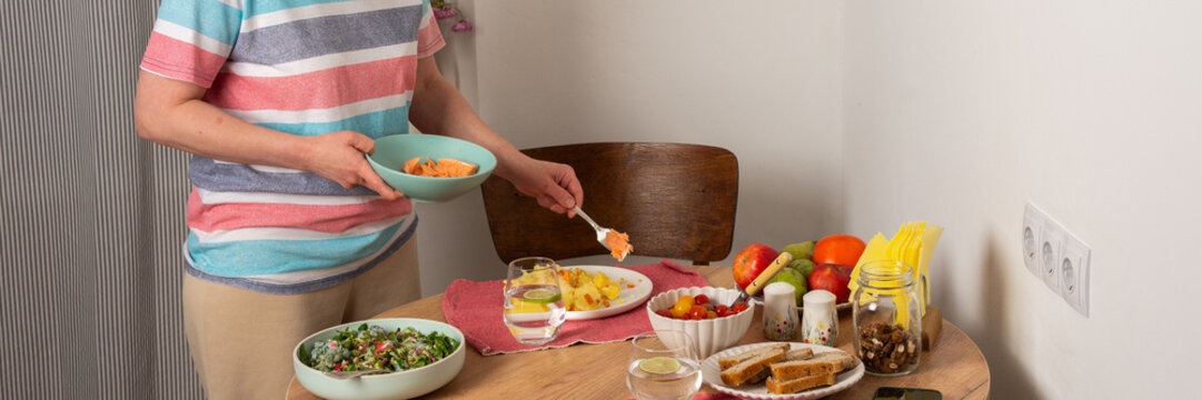 A 53-year-old woman is preparing dinner at home. She is setting the table while holding a pan with food. The setting is simple and warm, reflecting a typical evening meal time, banner