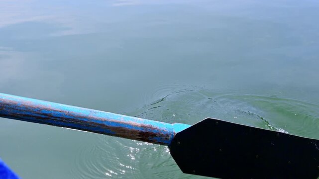 A close-up of a weathered oar slicing through calm waters of Naukuchiatal Lake, India, creates gentle ripples and droplets, reflecting sky on its smooth surface.