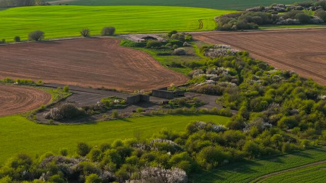 Concrete airfield remnants and bunker shelters surrounded by farmland and dense vegetation patches in countryside. Elevated drone view across abandoned World War II aviation infrastructure. Scenic