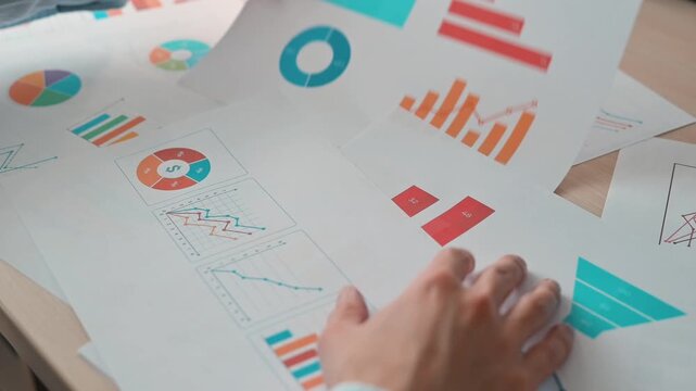 A person sorts through papers showing different charts and diagrams in an indoor setting. The individual, a young adult male, is focused on examining the information related to business data analysis