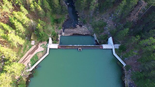 Aerial view of small forest dam with calm reservoir and controlled water drainage structure. Concrete spillway sits among dense trees creating quiet balanced man made liquid landscape.