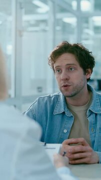 Young male patient sitting in clinic, giving clipboard with medical records to doctor and explaining his symptoms during consultation. Vertical over the shoulder shot