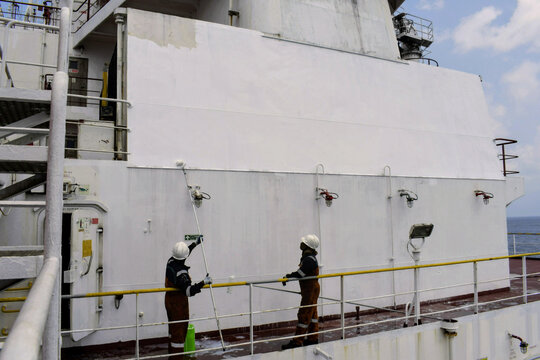 seafarers applying fresh paint on vessel superstructure using long rollers, showcasing routine maintenance, safety gear and teamwork at sea