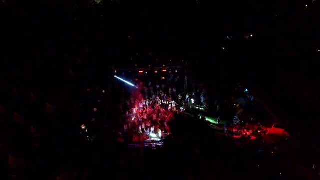 Drone aerial view captures outdoor night disco party crowd dancing around DJ stage. Energetic open air scene shows light beams spotlights pulsing wild music vibes useful lifestyle context.