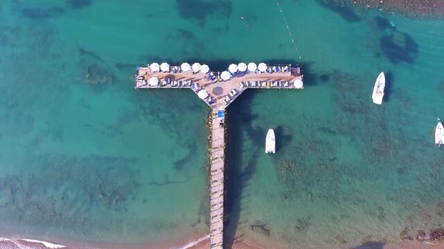 Aerial drone view of T shaped hotel pier extending into turquoise tropical sea with sun loungers. Luxury beach umbrellas line the deck as tourists enjoy calm summer vacation by ocean.