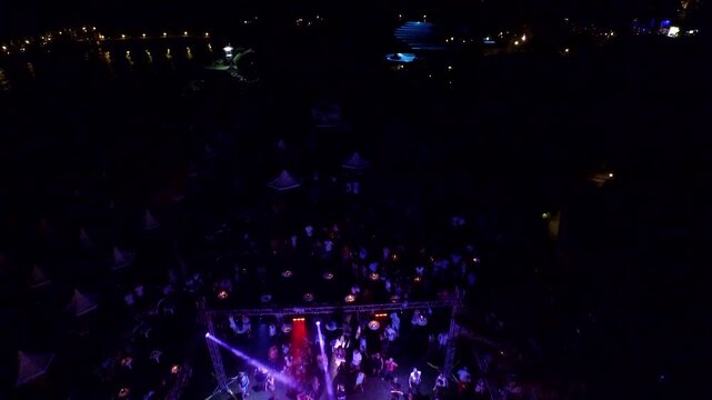 Drone aerial view captures outdoor night disco party crowd dancing around DJ stage. Energetic open air scene shows light beams spotlights pulsing wild music vibes useful lifestyle context.