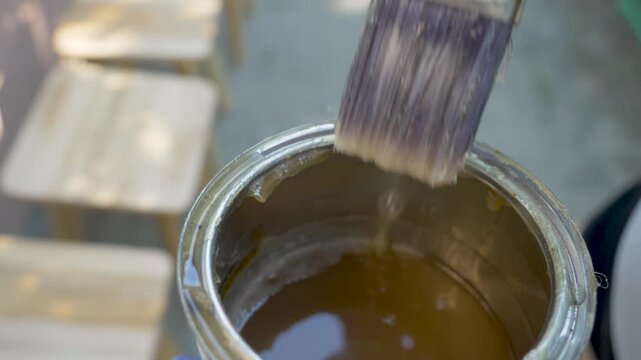 Brush submerges into varnish can in craftsman gloved hand. Tool soaks up coating, preparing for precise spread on wooden surface for protection and shine