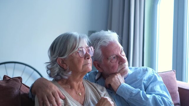 A loving elderly couple sits embraced on a comfortable sofa in their living room, looking contemplatively towards the window. This tender moment captures their lasting bond and shared thoughts 