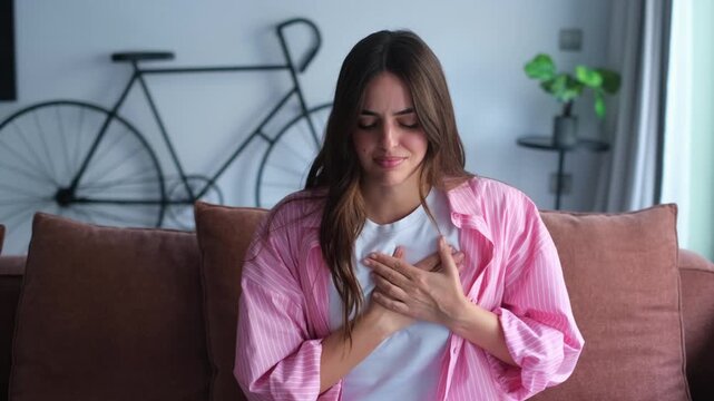 A concerned young woman sitting on a comfortable couch, clutching her chest and struggling with sudden pain or discomfort. She takes deep breaths, trying to find relief from the unsettling sensation.
