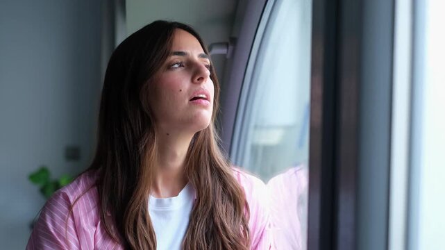A young woman looks out a window with a pensive expression, displaying feelings of loneliness, worry, and quiet introspection, perhaps reflecting on life or experiencing a moment of solitude.