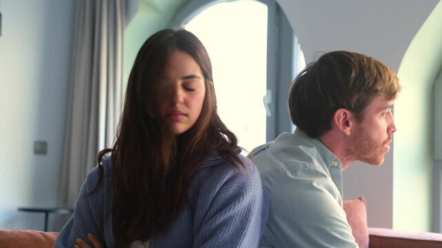 A young couple sits back-to-back on a sofa, deeply upset and avoiding eye contact after a disagreement. This video captures the emotional distance and sadness of a relationship crisis.