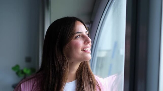 A beautiful young woman with a gentle smile gazes thoughtfully out a window, exuding happiness and contentment in the peaceful indoors of her home.