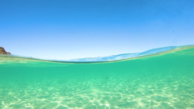 Slow motion split view showing the turquoise sea surface and the white sand beach above, with the crystal clear shallow seabed below at La Pelosa Stintino