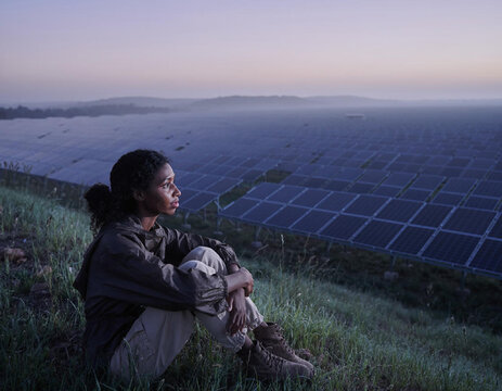 Woman sitting on green hill overlooking solar panels.