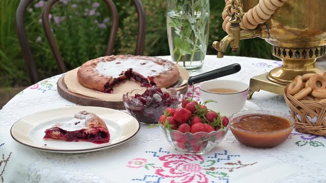 A rustic tea setting with golden samovar, fresh strawberries, jam, pie, and bagels arranged on an embroidered tablecloth outdoors.