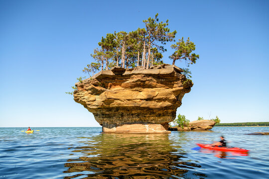 Kayakers paddle through the clear blue waters of Lake Huron surrounding Turnip Rock, a unique stack formation topped with a small grove of pine trees near Port Austin, Michigan.