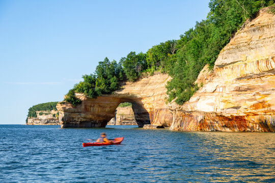 A kayaker glides across the deep blue water of Lake Superior toward Lovers Leap, a majestic natural sandstone arch along the colorful shoreline of Pictured Rocks National Lakeshore.