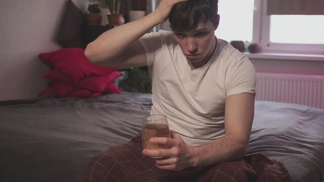 Young man suffering from strong headache or migraine sitting with glass of water in the kitchen, millennial guy feeling intoxication and pain touching aching head