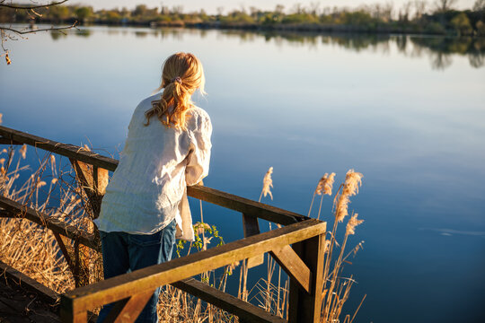 Woman standing on lakeside terrace enjoying view. Female person overlooking calm water from wooden deck. Serene moment in rural nature