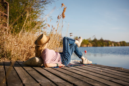 Relaxed woman with straw hat lying on wooden dock enjoying sun. Leisure time on jetty by water