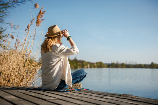 Solitary relaxation and mindfulness in nature. Woman with hat looking over peaceful lake from dock enjoying sun and fresh air outdoors