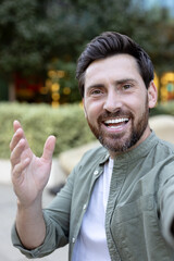 Naklejka premium Cheerful man with a beard and dark hair smiles broadly and waves his hand while capturing a selfie outdoors, embodying happiness, welcome, and positivity