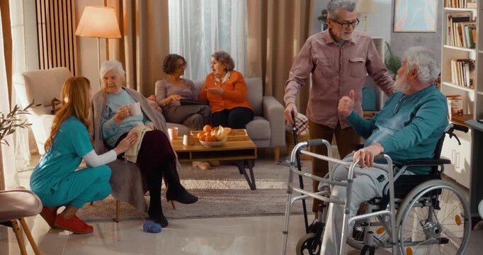 Wide view of seniors socializing in assisted living home. Caregiver supporting elderly woman while others relax, talk and spend time together, showing warm, everyday atmosphere and community life 