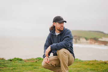 Man crouches near coastal grass, gazing thoughtfully © Reuben