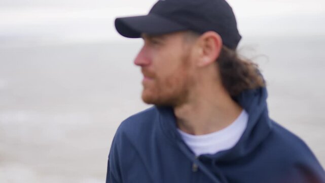 Man gazes at the ocean wearing a baseball cap and hooded jacket
