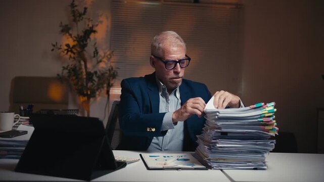Overwhelmed older male manager feeling stressed and frustrated with a huge stack of documents while working late at night in the office