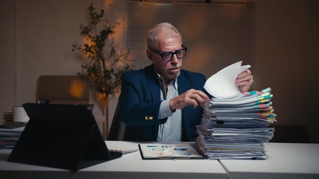Elderly manager sitting at his desk, overwhelmed by a large stack of documents. Stressed and tired man working late in a dark office