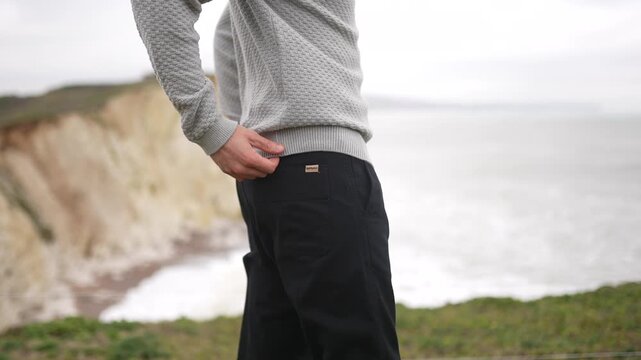 Man in sweater and dark pants stands overlooking dramatic coastal cliffs