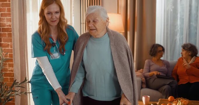 Kind caregiver helping elderly woman stand from armchair and walk with walker in assisted living home. Supportive moment of mobility, safety and independence indoors.