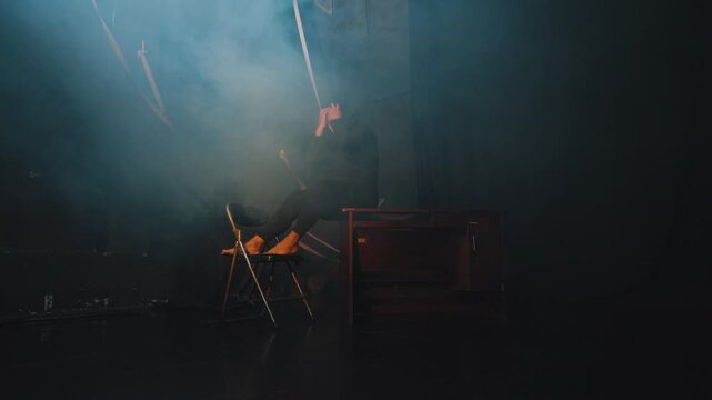 A distressed woman sits on a chair in a dark, smoky room, leaning forward with her head in her hands. The cinematic fog and lighting emphasize feelings of anxiety, grief, and mental struggle.