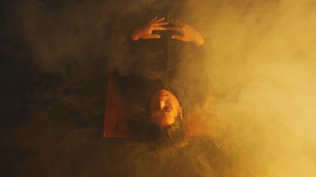 A dramatic top-down view of a woman lying motionless as heavy, vibrant yellow fog rolls over her face and body. The surreal aesthetic suggests a deep trance, dream state, or eerie slumber.
