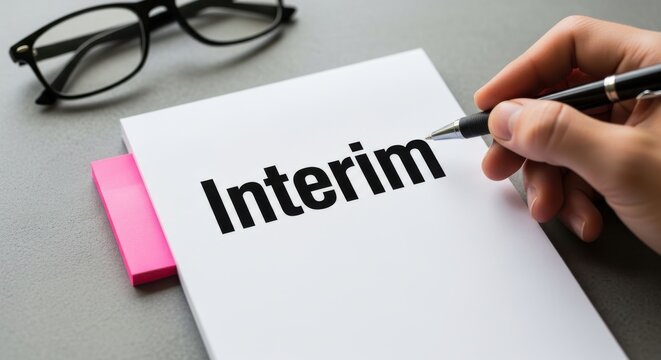 Interim business management concept, person holding pen over interim text on white paper, professional office workspace with glasses and sticky notes