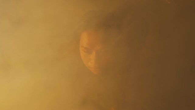 A woman's face slowly reveals itself from a thick cloud of yellow fog. She stares intensely before looking down with clasped hands, creating a suspenseful and mysterious atmosphere.