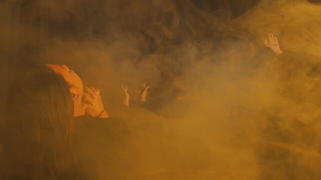 A woman with clasped hands watches as figures in the background perform expressive movements through thick yellow fog. The scene is highly atmospheric, dramatic, and artistically ambiguous.