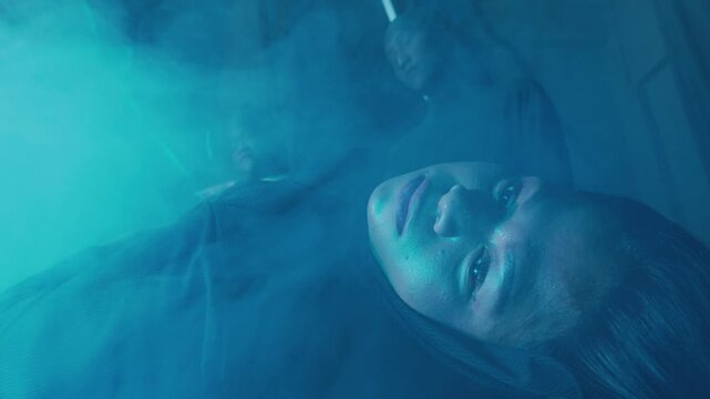 A haunting close-up of a woman lying on her back, eyes wide open in a frozen stare, while shadowy figures stand behind her in thick cyan fog. Depicts sleep paralysis or a surreal nightmare.
