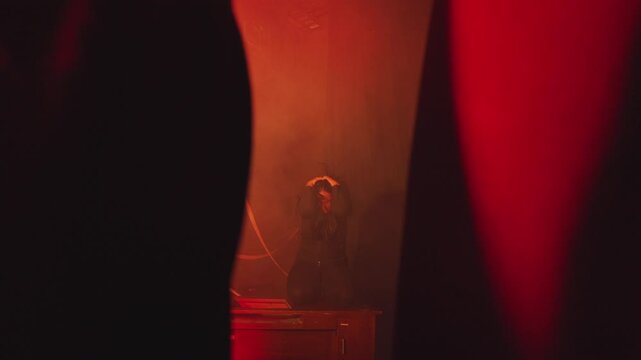 Wide shot of a woman kneeling on a wooden prop in a dark room filled with red smoke. Foreground silhouettes frame her as she holds her head in a dramatic, theatrical display of emotional suffering.