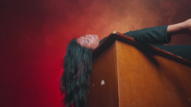 Beautifully lit conceptual footage of a woman lying on her back across a wooden desk. Deep red and orange smoke fills the background, symbolizing burnout, emotional exhaustion, or a dramatic defeat.