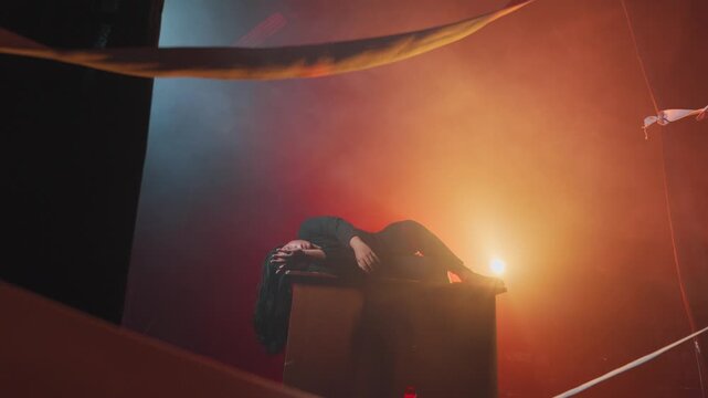 An artistic composition featuring a woman collapsed on a wooden structure. Surrounded by intense red and orange smoke, she raises an arm weakly, portraying themes of burnout, failure, or deep tragedy.