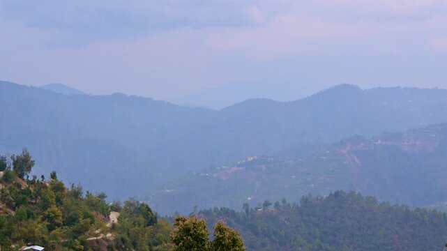 Time-lapse view of Mukteshwar, Uttarakhand, India, capturing layered mountain ridges bathed in mist, soft sun rays streaming through clouds, illuminating scattered hillside homes and winding trails.