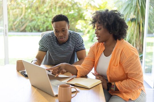 African American friends sitting at table by sliding glass door, pointing at laptop, orange shirt