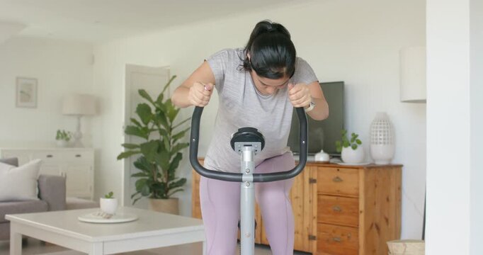 Woman wearing leggings, headphones bringing hands to curved handlebars, pedaling at home for cardio