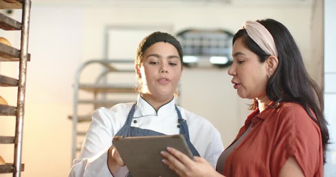 Diverse female bakery staff manager pointing at tablet, chef in coat tapping to confirm bread batch