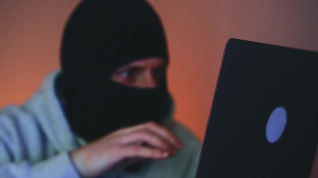 Close-up of a male hacker working on a laptop in a dark environment. The man is engaged in illegal cyber activity, hacking a server and stealing sensitive data. The scene conveys secrecy, danger, cybe